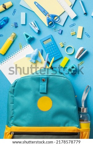 Back to school concept. Top view vertical photo of school supplies flying out of blue schoolbag on isolated blue background