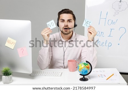 Employee confused operator business man in set microphone headset for helpline assistance work at call center office desk pc computer hold YES or NO symbol card make choice isolated on grey background
