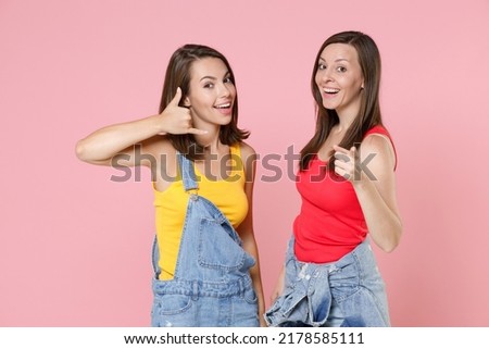 Two cheerful excited young women friends 20s in casual denim clothes doing phone gesture says call me back pointing index finger on camera isolated on pastel pink colour background, studio portrait