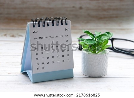 July 2023 white desk calendar on wooden table with plant and glasses background. Copy space