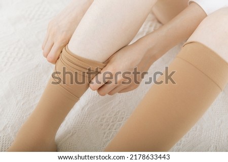 Beautiful long female legs in stockings. Girl putting on stockings at home in a white room. Beige knee socks. Varicose veins prevention. Woman body in underwear.