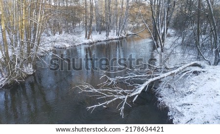 The river flows through the forest. In late fall in the forest the trees stand leafless and reflected in the water of the river After the snowfall, snow lies on the river banks and on the trees. Sunny Royalty-Free Stock Photo #2178634421