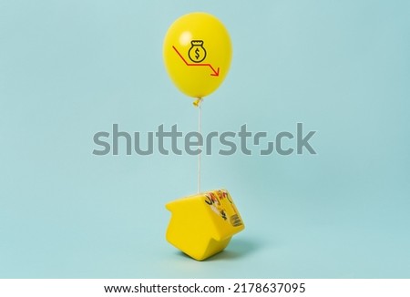Inflation symbol.
A yellow house-shaped piggy bank lifted by a yellow balloon with a depreciating dollar sum.  Yellow Money box at blue background
