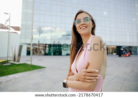 Attractive stylish european woman in sunglasses and pink summer dress posing on city background with modern buildings in sunny day.