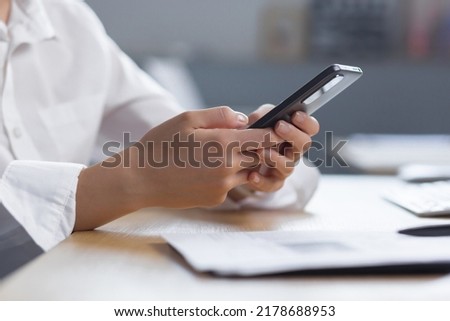 Close-up photo. The hands of a young woman in a white shirt are holding a mobile phone, typing a message at the table.