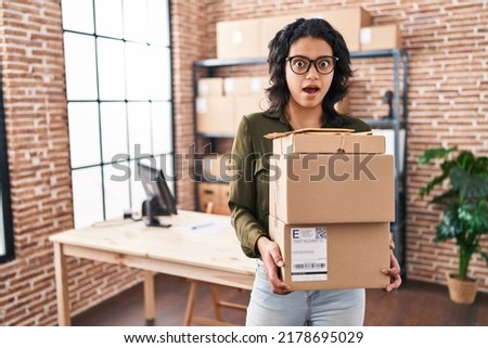 Hispanic woman with dark hair working at small business ecommerce holding boxes in shock face, looking skeptical and sarcastic, surprised with open mouth 