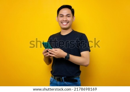Cheerful young Asian man in casual t-shirt using mobile phone and standing confident looking at camera isolated on yellow background. People lifestyle concept