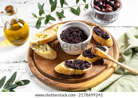 Olive pate in bowl and spreader on bread slices on wooden cutting board.  Royalty-Free Stock Photo #2178699241