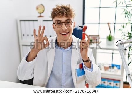 Young caucasian doctor man holding ear otoscope at the clinic doing ok sign with fingers, smiling friendly gesturing excellent symbol 
