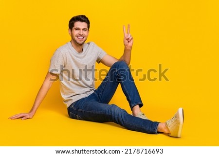 Full body photo of nice brunet millennial guy sit show v-sign wear t-shirt jeans footwear isolated on yellow background