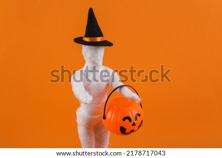 Trick or treat. Mummy doll wrapped in bandages in a witch's hat with a jack lantern candy bucket on orange background. Halloween minimal still life. creative layout