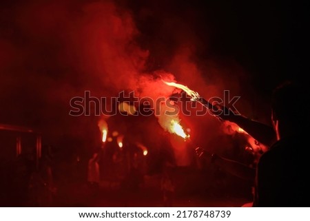 Red bengal torch flame burning and creating particles of fire, light and smoke in the dark, high contrast pyrotechnics shot. Celebrating winning, football hooligans concept. Royalty-Free Stock Photo #2178748739