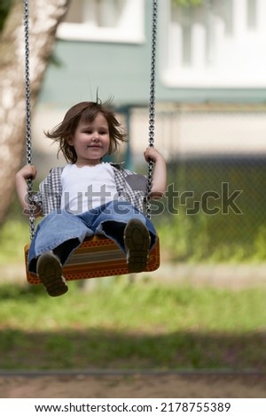 A smiling little girl is swinging on a playground swing in the yard of the house. Her hair is flying. Selective focus.                              