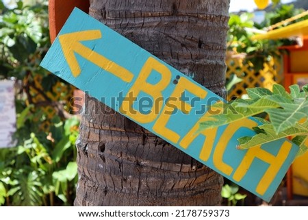 Closeup of a blue sign on a tree with yellow text with the words beach and an arrow to the left showing the direction