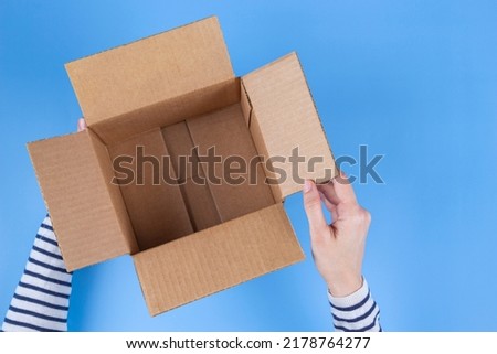 Woman hands holding open empty brown cardboard box on light blue background. Top view