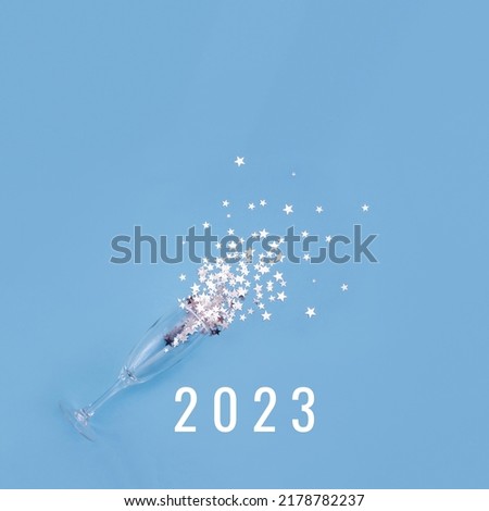 Champagne glass on blue background glass surrounded by silver star confetti and numbers 2023 . New Year celebration or party concept. Selective focus, copy space