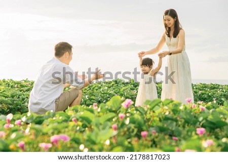 A father was stretching his hands open to welcome her daughter who was learning to walk towards him along the flowers road