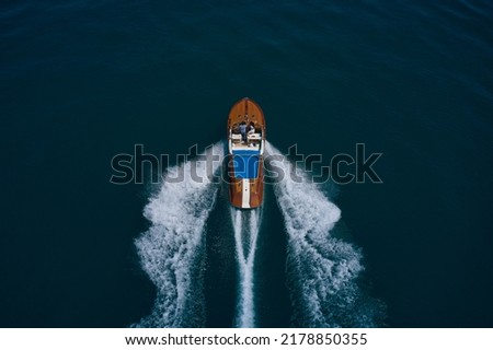 Wooden expensive italian boat with people fast movement on the water top view. High speed open modern wooden boat moving fast on dark water aerial view. Royalty-Free Stock Photo #2178850355