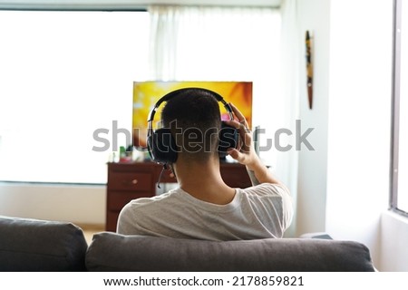 person from the back sitting on a sofa in the living room of a house, using wireless headphones to listen to music in front of television, technology and lifestyle with hobbies