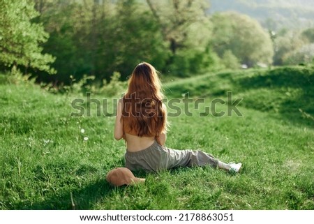Top view of a woman in an orange top and green pants sitting on the summer green grass with her back to the camera with her phone, a young freelance student's concept of work and leisure Royalty-Free Stock Photo #2178863051