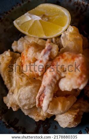 Deep-fried tempura shrimp and squid with a lemon wedge next to it. The food is in a blue, deep, ceramic plate. The plate stands on a wooden background, next to it there is a glass of white wine, a pep