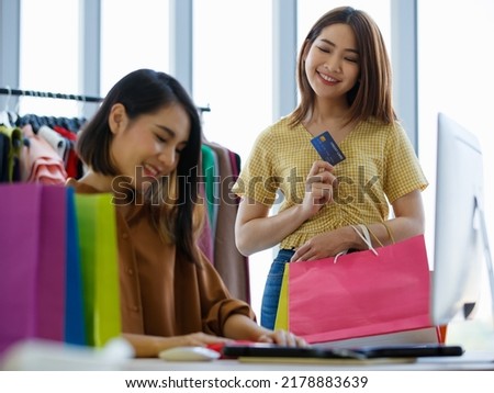 Asian female seller smiling and putting garment in paper bag for happy woman with credit card in shop Royalty-Free Stock Photo #2178883639