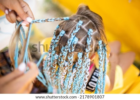 A little waiting tanned girl in a bright yellow summer suit, weaves colored pink-blue African braids in her dark wet thick hair on a hot sunny sea day Royalty-Free Stock Photo #2178901807