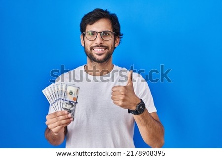 Handsome latin man holding dollars banknotes smiling happy and positive, thumb up doing excellent and approval sign 