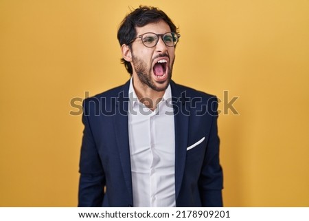 Handsome latin man standing over yellow background angry and mad screaming frustrated and furious, shouting with anger. rage and aggressive concept. 