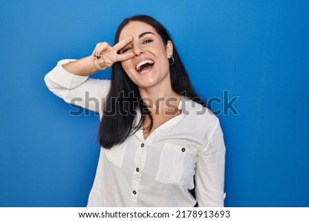 Young hispanic woman standing over blue background doing peace symbol with fingers over face, smiling cheerful showing victory 