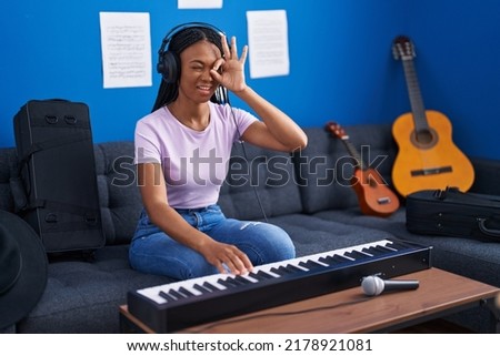 African american woman with braids playing piano keyboard at music studio smiling happy doing ok sign with hand on eye looking through fingers 