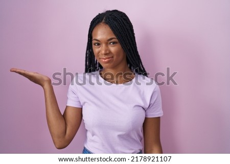 African american woman with braids standing over pink background smiling cheerful presenting and pointing with palm of hand looking at the camera. 