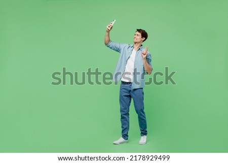 Full size side view fun young brunet man 20s wears blue shirt get video call use mobile cell phone conducting pleasant conversation show victory sign isolated on plain green background studio portrait