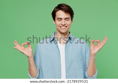 Spiritual tranquil happy young brunet man 20s years old wears blue shirt hold spreading hands in yoga om aum gesture relax meditate try to calm down isolated on plain green background studio portrait