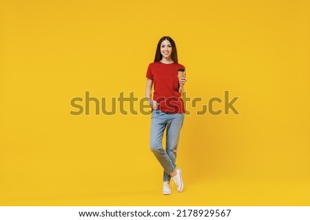 Full size body length young brunette woman 20s wears basic red t-shirt hold takeaway delivery craft paper brown cup coffee to go isolated on yellow background studio portrait. People emotions concept
