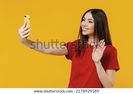 Beautiful fun young brunette woman 20s wears basic red t-shirt doing selfie shot on mobile cell phone post photo on social network meet greet waving hand isolated on yellow background studio portrait.