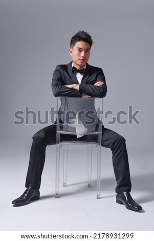Handsome fashion model.
elegant man wear formal black suit with bow tie sitting chair on gray background 


