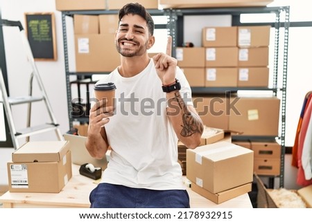 Young hispanic man working at small business ecommerce drinking a coffee surprised with an idea or question pointing finger with happy face, number one 