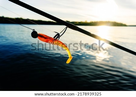 A fragment of a spinning rod and a silicone bait against the background of the river and the sun's rays. Fishing for predatory fish in the summer. Foreground Royalty-Free Stock Photo #2178939325