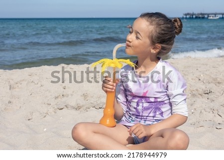 A little girl drinks juice while sitting on the sand by the sea on a sunny summer day.