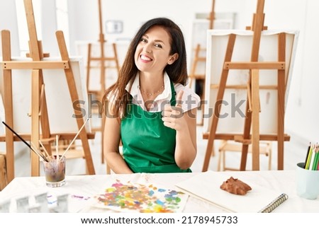 Beautiful caucasian woman at art studio smiling happy and positive, thumb up doing excellent and approval sign 