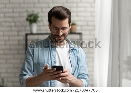 Happy millennial caucasian man in glasses look at smartphone screen browse wireless internet on gadget, smiling young male text message on modern cellphone at home, communication concept Royalty-Free Stock Photo #2178947705