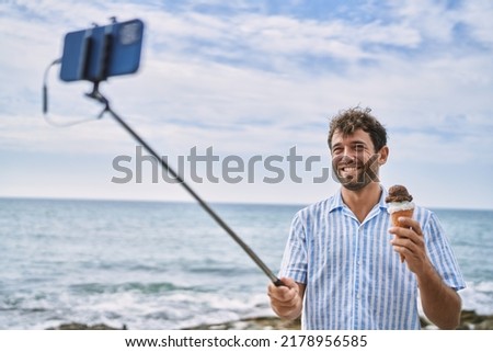 Young hispanic man eating ice cream making selfie by the smartphone at the beach.
