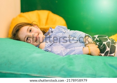 Happy smiling girl lying on green mattress in kindergarten with multi colored interior and looking at camera carefully