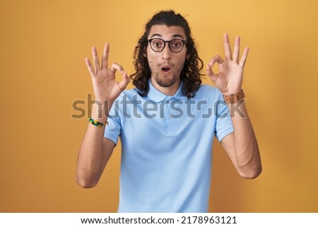 Young hispanic man standing over yellow background looking surprised and shocked doing ok approval symbol with fingers. crazy expression 