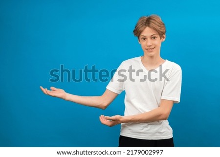 Young teen boy points palms aside isolated on blue. Friendly well-looking boy paying your attention at copy space, best deal, sale, presenting novelty