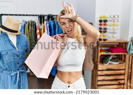 Young caucasian woman holding shopping bags at retail shop making fun of people with fingers on forehead doing loser gesture mocking and insulting. 