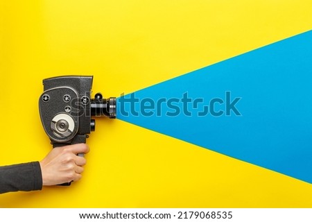 Female hand holding old style film movie camera on a yellow background with blue ray coming out of the camera,  imitating shooting process. Ukrainian flag colors. Film making industry concept