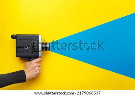 Female hand holding old style film movie camera on a yellow background with blue ray coming out of the camera,  imitating shooting process. Ukrainian flag colors. Film making industry concept
