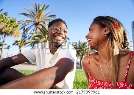 Cheerful young African couple laughing and having fun on their first date in the park in a romantic outdoor lovers scene, sitting on the grass talking Royalty-Free Stock Photo #2179099961